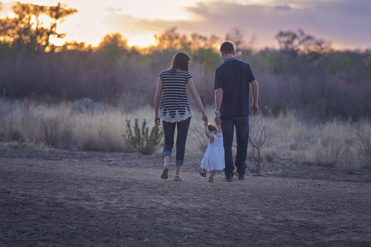 Two parents walking away from the camera at dusk, holding the hands of a small child between them.